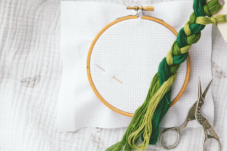 Embroidery hoop with white fabric, two needles, green thread braid and scissors on soft textile,...