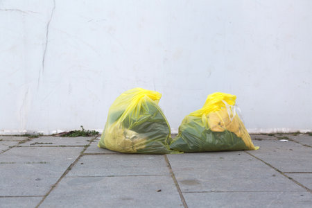 Two yellow garbage bags full of waste standing on gray pavement near a white wall, concept of...