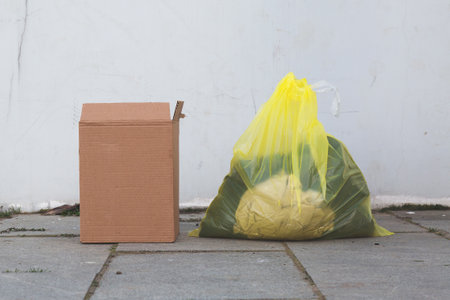 A yellow garbage bag filled with waste and a damaged cardboard box placed on a sidewalk against a white wall, representing waste management and recycling effortsの写真素材