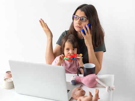 Busy mother working remotely with child on lap, talking on smartphone, surrounded by toys and laptop, concept of parentingの写真素材
