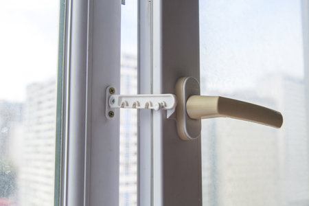 Plastic safety window limiter with beige handle installed on white frame, partially open window with...