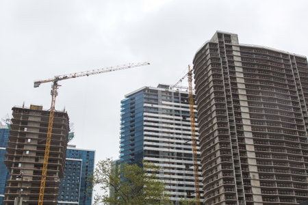 Unfinished modern high-rise buildings with exposed concrete and yellow tower cranes under cloudy sky, urban area development in progress, concept of architectureの写真素材
