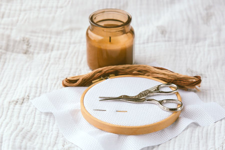 Wooden embroidery hoop on white canvas with metal scissors, brown floss and candle on soft fabric...