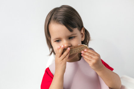 child in a red shirt and pink bib eating a slice of bread, focusing intently, illustrating a moment of simple and wholesome mealtime at homeの写真素材