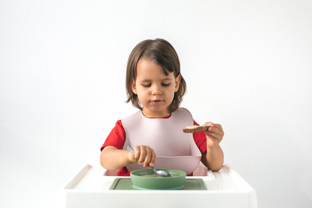 Little girl with brown hair sitting in a high chair, wearing a pink bib and red shirt, eating porridge with a spoon and holding a piece of bread against a plain white backgroundの写真素材