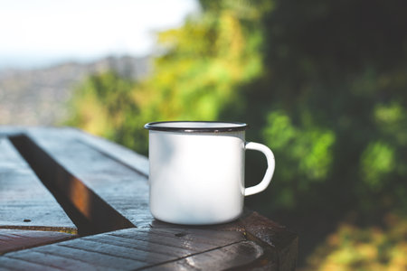 White enamel mug on a wooden table outdoors with green forest background, morning sunlight and...