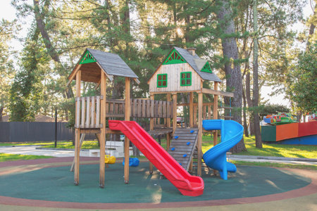 A wooden empty playground structure with slides and swings surrounded by trees in a sunny park, featuring red and blue slides and green decorative details, concept of childhood and outdoor playの写真素材