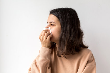 Woman using a tissue to wipe her nose, wearing a beige sweater, illustrating symptoms of a cold, allergies, or respiratory illness, isolated against a white backgroundの写真素材