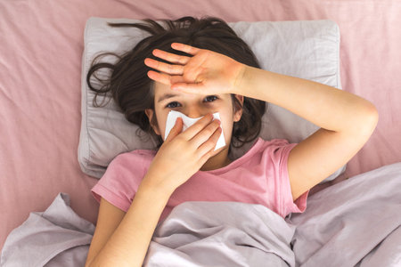 A sick girl lying in bed with a tissue covering her nose, wearing a pink t-shirt, and holding her hand to her forehead, looking unwell. Concept of illness and recoveryの写真素材