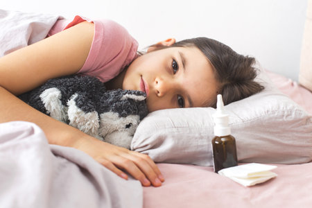 Young girl child lying in bed with a tired expression, hugging a stuffed animal, a nasal spray and tissues nearby, illustrating illness or recovery from a cold, isolated in a cozy environmentの写真素材