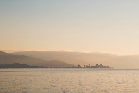 A distant view of Batumi city at sunset, with its modern skyline silhouetted against the hazy mountains and calm sea, under a warm golden sky. Concept of urban landscapes and travelの写真素材