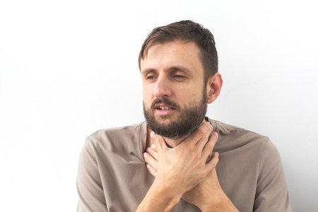 Bearded man in a beige T-shirt holding his throat with both hands, looking unwell. His facial...