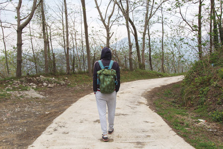 Person in hoodie walking through forest on a paved trail with a green backpack, early spring scenery...