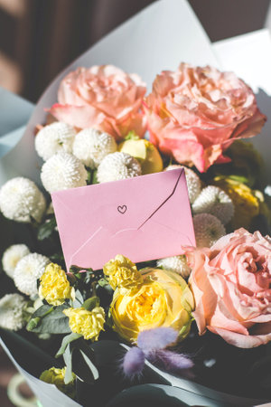 Pink envelope with a small heart symbol placed among fresh flowers, including roses and chrysanthemums, symbolizing love, romance, and special occasions concept and valentines day, vertical photoの写真素材