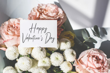 Stylish minimalist bouquet for Valentine's Day with pastel roses, white chrysanthemums, and eucalyptus, complemented by a handwritten greeting card with text Happy Valentine's Day, in natural sunlightの写真素材