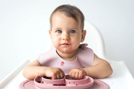 Adorable baby with blue eyes sitting in a high chair, wearing a pink bib, and eating messy food....