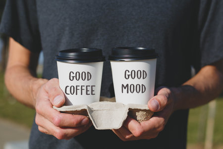 close-up of two takeaway coffee cups held in a cardboard holder. One cup reads Good Coffee, other Good Mood. The person wears a dark T-shirt, blurred outdoor. concept of love addiction to coffee.の写真素材
