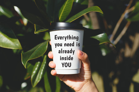 Hand holding a takeaway coffee cup with motivational quote text Everything you need is already inside you against green leaves background in sunlight, concept of inspirationの写真素材