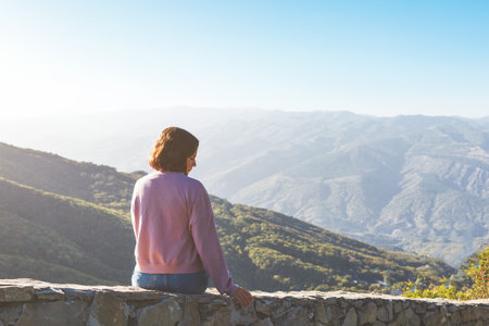 young female tourist woman in pink sweater sits in the mountains and admires the beautiful mountain...