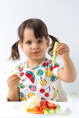 Little girl with pigtails sitting in a high chair, eating fresh vegetables and holding a cucumber stick, smiling playfully. Healthy eating habits for children.の写真素材