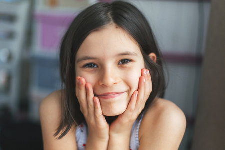Cheerful 7-8 year old girl with long dark hair smiling with hands on cheeks in cozy home...