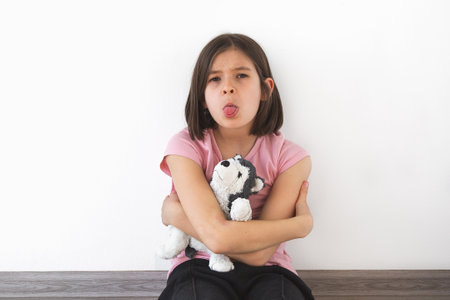 child girl 7-8 years in pink t-shirt sits against white wall, hugging plush toy and sticking out her...