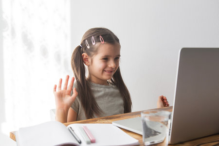 A girl 6 years old in front of a laptop monitor waving hand to someone. Online education and online...
