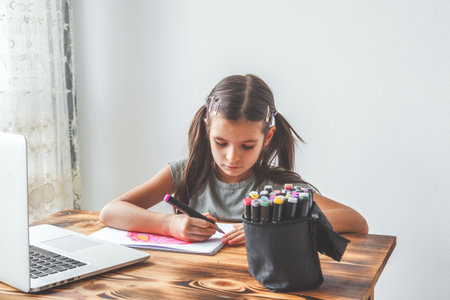 child girl 6-7 years old preschooler schoolgirl sits at a table in front of a laptop and draws with...