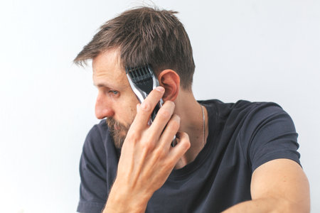 Young man aged 30 35 cutting his hair with electric clipper at home, focused on trimming around ear, dressed casually, white background, self-grooming concept of lifestyleの写真素材