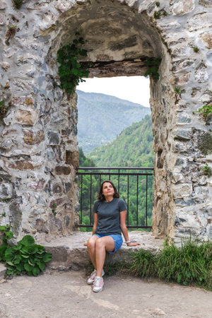Vertical photo of young woman sitting in stone ancient mountain fortress, surrounded by nature looking up thoughtfully, with lush green valley hills, concept of travel, historical explorationの写真素材