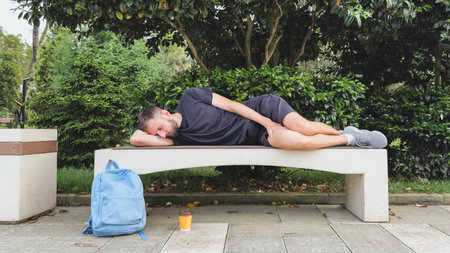 Young man in casual sportswear sleeping on modern stone bench in green city park, next to blue...