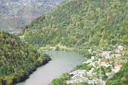 A serene view of a small Georgian village nestled in the green mountainous region of Adjara, with a calm river flowing through the valley and lush forests surrounding the settlementの写真素材