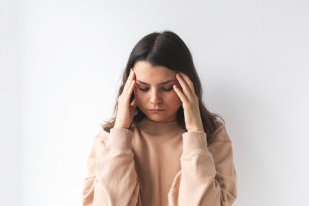 A young woman holding her head in discomfort, possibly experiencing stress, headache, or fatigue, with a neutral background emphasizing her emotionsの写真素材