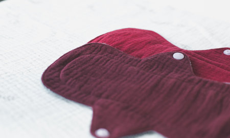 Close-up of reusable cloth muslin menstrual pads in burgundy color, placed on a white textured...