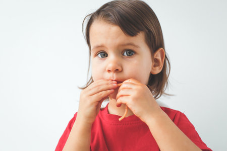 Little child eating spaghetti with hands, wearing a red shirt, looking thoughtful and curious,...