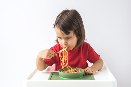 A 2.5-year-old girl in a red shirt sitting in a high chair, enthusiastically eating spaghetti with tomato sauce from a green bowl on white background. Concept of childhood and mealsの写真素材
