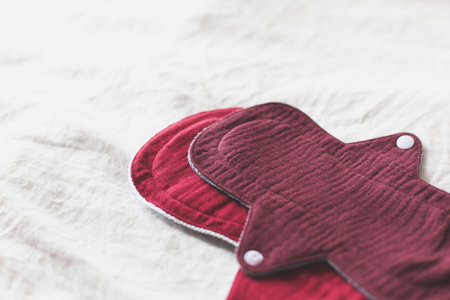Reusable cloth menstrual pads made of soft, deep red fabric with snap closures, on white textured surface, symbolizing sustainable and eco-friendly alternatives for personal hygiene zero-waste livingの写真素材