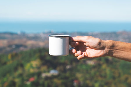 Hand holding a white enamel mug against a blurred scenic background of green hills and the sea under a clear blue sky, evoking simplicity and outdoor lifestyleの写真素材