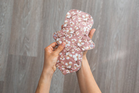 Reusable cloth menstrual pad with a cherry blossom pattern in pink tones held by two hands on a wooden floor background concept of sustainabilityの写真素材