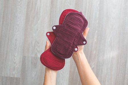 Reusable cloth menstrual pads in burgundy color held by a woman's hands against a neutral background. Concept of women's health, sustainability, and eco-friendly lifestyleの写真素材