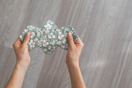 Reusable cloth menstrual pad with a floral pattern held by two hands on a wooden floor background concept of sustainabilityの写真素材