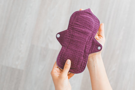 A reusable menstrual pad in dark purple fabric held in hands against a light wooden background, promoting sustainable and eco-friendly menstrual care, concept of health and sustainabilityの写真素材