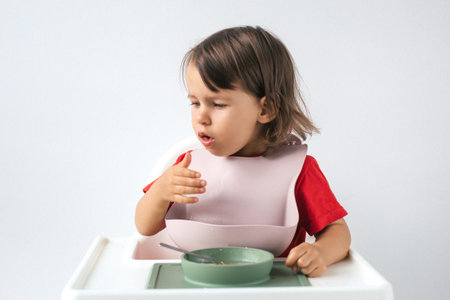 Little girl sitting in high chair, wearing pink bib and red shirt, coughing while eating. holds a spoon in one hand, with food on her lips, and touches her chest, showing discomfort, choking on mealの写真素材