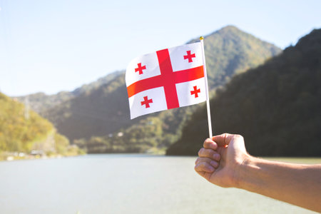 A hand holding the flag of Georgia against the backdrop of the picturesque mountainous landscape of Adjara, with green hills and a serene lake in the background. Concept of patriotism and travelの写真素材