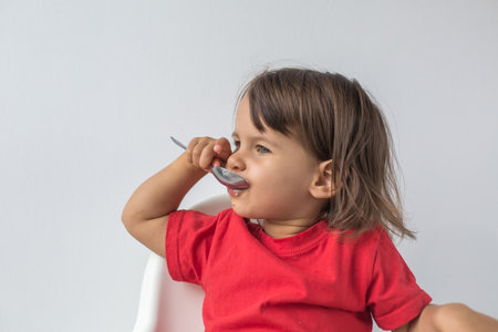 Little girl with dark hair wearing a red shirt sitting in a high chair, happily eating soup with a spoon from a green bowl. Concept of childhood, independence, and healthy eatingの写真素材