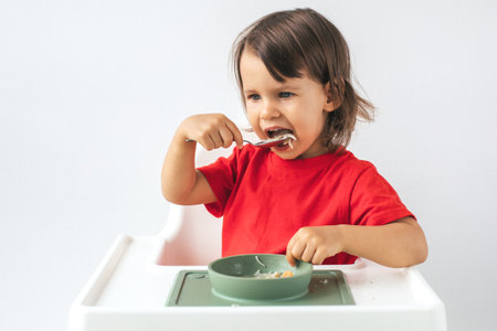 Little child girl sitting in a high chair on white background, wearing a red shirt, eating noodles with a spoon from a green bowl. She looks focused as she learns to feed herself independentlyの写真素材