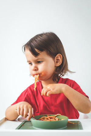Child eating spaghetti with tomato sauce, sitting in high chair and making mess. The toddler enjoys the meal with a playful expression, highlighting childhood, food, and family life concept, verticalの写真素材