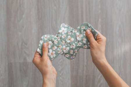 Hands holding a reusable cloth menstrual pad with a floral print in pastel green, highlighting eco-friendly and sustainable period care on a wooden background, concept of menstruationの写真素材