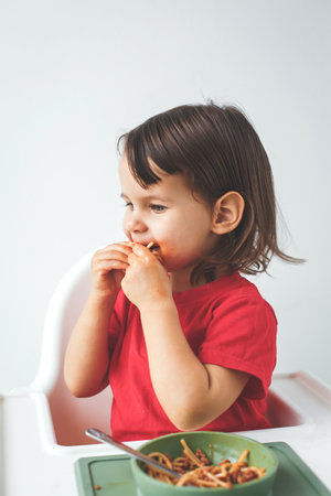 A toddler girl in a red shirt enjoys eating spaghetti with hands, sitting in a high chair, with a green bowl and a fork on the tray, against a clean white background in kitchen, concept of baby foodの写真素材