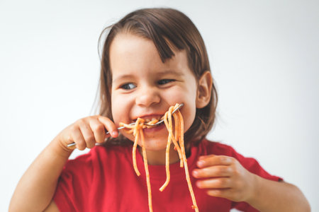 Happy child girl in a red shirt eating spaghetti with a fork, smiling and enjoying a playful and messy moment, close-up portrait with a white background, concept of childhoodの写真素材